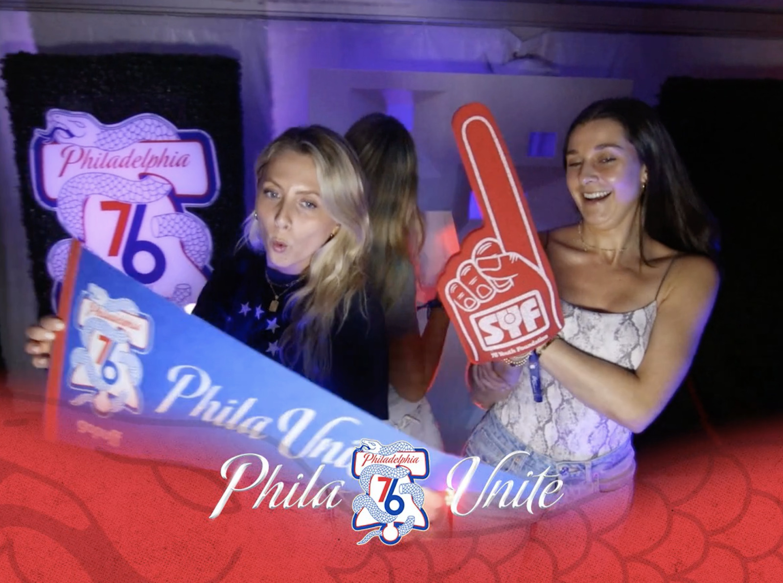 Dos chicas de fiesta. Una sostiene una bandera de los Philadelphia 76ers y la otra un dedo de espuma.