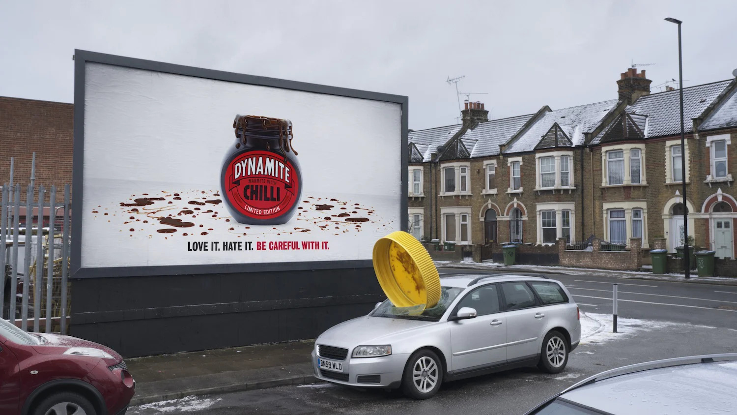 Billboard of an empty Dynamite Chilli jar with a missing lid. In real life, a car sits in front of the billboard with a large yellow lid stuck in its hood