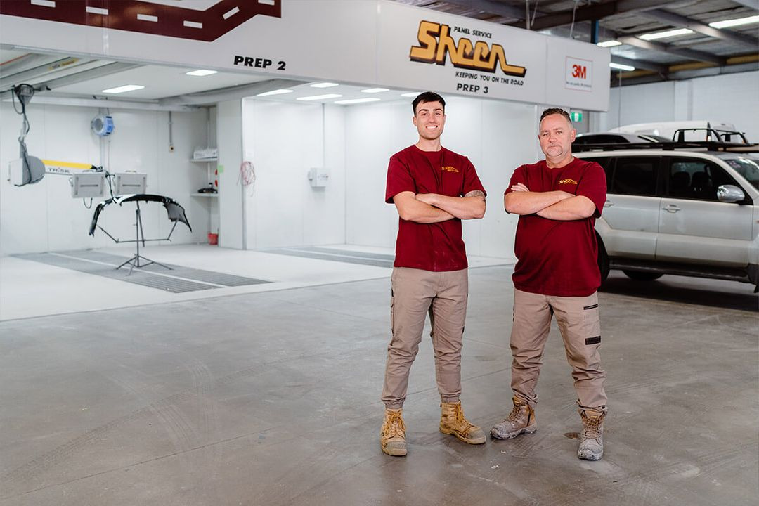 The Sheen Panel Services team is wearing uniforms and is in the garage where they repair damaged vehicles.