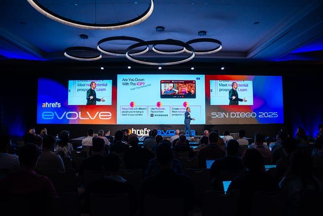 A speaker presents on a stage to an audience, with three large screens showing presentations from Ahrefs Evolve San Diego 2025.